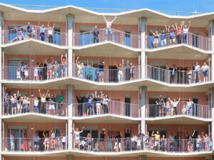 Das Bild zeigt vier Stockwerke des Laubengangs des Hauses. Auf jedem Stock stehen zwischen zehn und zwandig Personen - alles Bewohner_innen von Vis-à-Wien. Ein Großteil der Personen springen in die Luft und Strecken die Arme in die Höhe