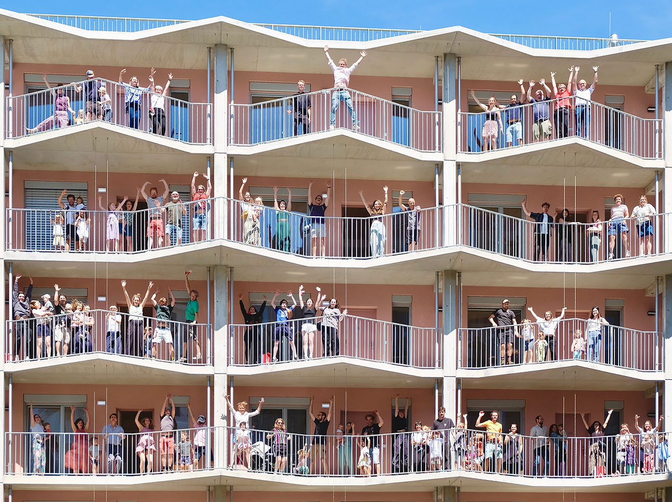Das Bild zeigt vier Stockwerke des Laubengangs des Hauses. Auf jedem Stock stehen zwischen zehn und zwandig Personen - alles Bewohner_innen von Vis-à-Wien. Ein Großteil der Personen springen in die Luft und Strecken die Arme in die Höhe