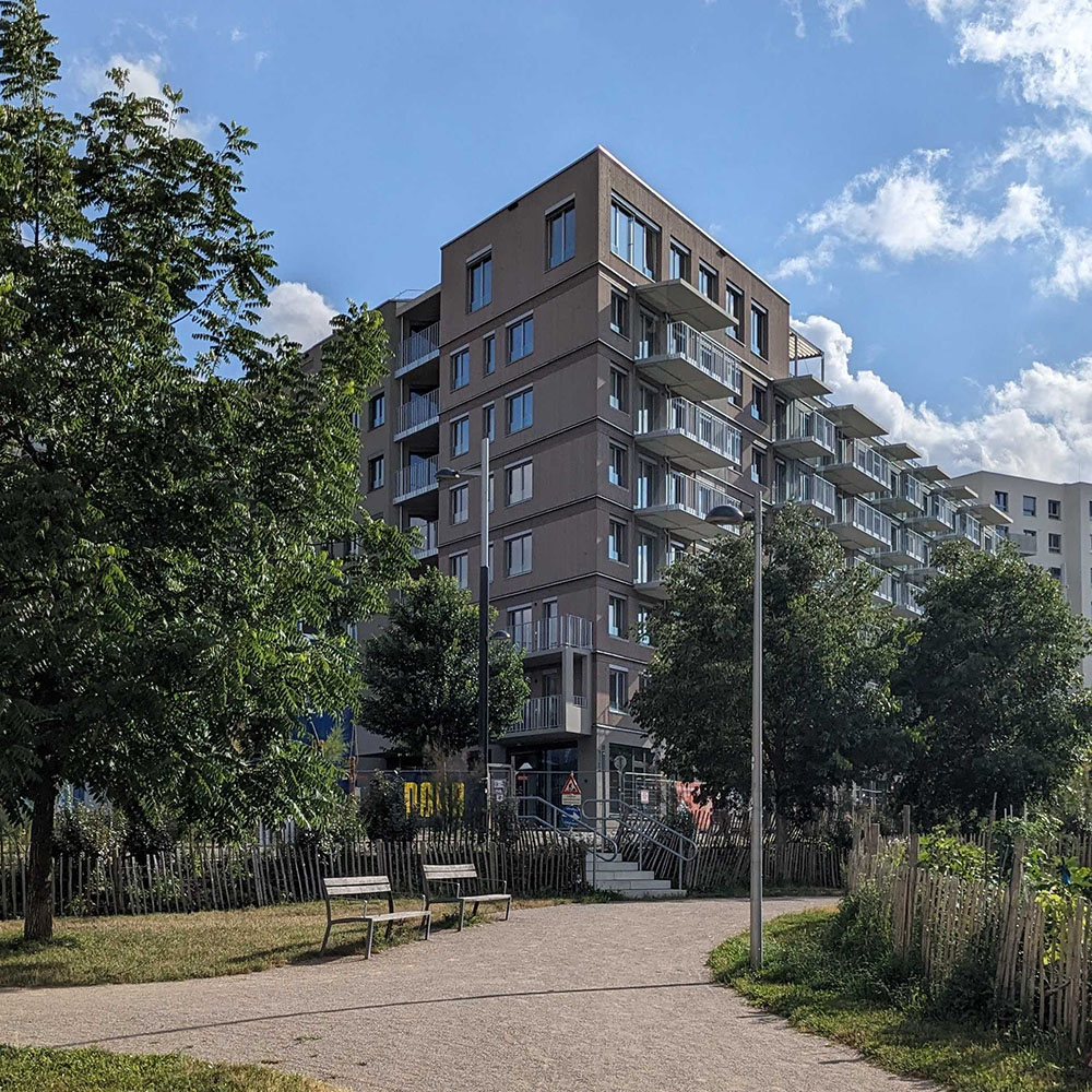 Foto vom Zelman-Park aus vom Nordost-Eck des Gebäudes. Im Vordergrund Bäume des Parks und dazwischen Stufen, die auf das höhere Niveau des Gebäudes hinaufführen.